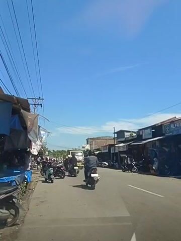 The video captures a street scene on a clear, sunny day. A paved road runs through the center, with lanes marked by white lines. On the left side of the road, several market stalls are set up, covered by blue and grey tarps. Various items, possibly goods for sale, are visible beneath the tarps, and a blue tarp is prominent in the foreground. Several motorcycles are parked along the side of the road near the stalls.

In the middle of the road, a yellow truck is moving away from the camera. A man is visible driving the truck. Several other motorcycles are also on the road, moving in the same direction as the truck. One motorcycle with a rider is closest to the camera. On the right side of the road, more market stalls or shops are lined up, with a sign that partially reads "ALFA." Motorcycles are parked in front of these establishments as well.

Overhead, a network of power lines stretches across the sky, supported by poles. The sky is a vibrant blue with minimal clouds. The overall atmosphere appears to be that of a bustling roadside market or town. The camera is positioned as if from inside a vehicle, looking forward.