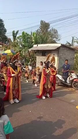This is a vertical, low-resolution, handheld video clip shot outdoors during the daytime. The scene depicts a parade or procession. In the foreground, two individuals are walking forward, dressed in elaborate, traditional costumes. They are wearing red and gold garments with ornate headpieces that feature feathery, fan-like structures. Both are wearing hijabs. To their right, behind them, a group of children, also in traditional costumes, are walking. These costumes are smaller versions of the adult outfits, featuring red and gold elements, and some children have similar headpieces.

To the right of the main procession, a red and white motorcycle is parked. A man in a blue shirt is standing next to it, looking towards the parade. In the background, there are several small buildings, some with corrugated metal roofs. There are trees and vegetation, including banana trees, suggesting a rural or semi-rural setting. The ground is paved with asphalt. The sky is visible and appears clear and bright. The overall atmosphere suggests a cultural event or celebration.
