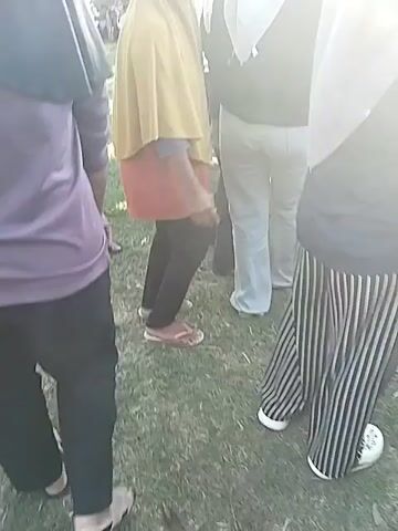 The video shows a crowd of people gathered in front of a stage, likely attending a concert or event. Most of the people in the foreground are wearing hijabs. The stage is set up with scaffolding, speakers, and what appears to be a large flag. The sun is bright, creating a somewhat overexposed and hazy view of the scene. Some individuals are holding drinks. The camera angle is from within the crowd, looking towards the stage.