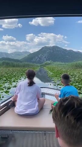 A video shows a view from the back of a motorboat as it travels down a narrow canal filled with lily pads and reeds. Two children are seated facing forward in the boat, looking at the scenery. In the foreground, the back of a third person's head is visible, also facing forward. The boat is moving through a lush green landscape with mountains in the background under a cloudy blue sky.