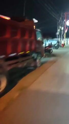 The video appears to be filmed at night, possibly from a moving vehicle or motorcycle, given the motion blur and perspective. The scene depicts a street with several motorcycles parked and people gathered, suggesting a busy area like a gas station or a small market. There is a prominent red and white structure on the left side of the frame, likely a building or shop, illuminated by bright lights. Further down the street, more lights are visible, possibly from other businesses or streetlights. The overall atmosphere seems to be that of a lively commercial or social hub during the evening.