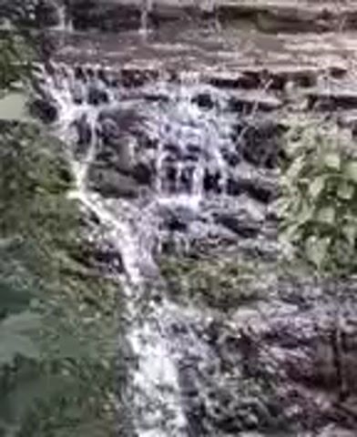 The video shows a close-up view of a waterfall cascading over rocks. The water is white and frothy as it flows downwards. Lush green foliage is visible on either side of the waterfall, suggesting a natural, outdoor setting. The water appears to be flowing steadily. The camera remains relatively still throughout the short clip, focusing on the movement of the water and the texture of the rocks.
