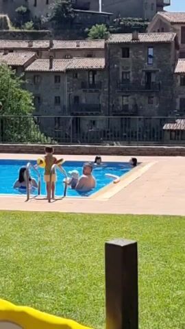The video shows a group of people enjoying a sunny day at an outdoor swimming pool. In the foreground, green grass and a tiled patio area are visible. The pool itself is bright blue and appears to be in use. Several people are in the water, including a man who is standing, and a child wearing yellow armbands. Others are sitting or lying on the grass around the pool. Some individuals are relaxing under beach umbrellas. In the background, traditional stone buildings with tiled roofs are visible, suggesting a village or historical setting. A tall tower, possibly part of a church, rises above the buildings. The overall atmosphere is one of leisure and recreation on a warm, clear day.