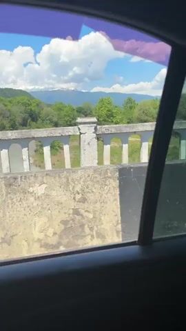 The video shows a view from the window of a moving car in Primorskoe, Abkhazia, on a bright, sunny day. The scene outside is dominated by lush green trees and vegetation, suggesting a natural or semi-rural environment.

At the beginning of the clip, a concrete barrier with white balustrades is visible close to the car, with a dense array of green trees and foliage behind it. As the car continues to move, the view shifts to a landscape featuring a metal guardrail alongside the road. Beyond the guardrail, there is a grassy area leading to more green trees under a clear, bright sky. The camera remains static inside the car, capturing the passing scenery through the window, which appears to have a slight tint. No text is visible in the video.