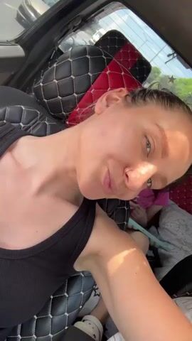 The video shows a woman in a moving vehicle, likely a car, taking a selfie. She is wearing a black tank top, has a nose ring, and her hair is pulled back in a bun.  She looks at the camera and smiles, and is seated in the front passenger seat, next to what appears to be a black seat cover with red accents. A young girl in a pink shirt is in the backseat, occupied with what appears to be a book or tablet. Other vehicles are visible through the window. The woman is speaking to someone but the audio is unclear.