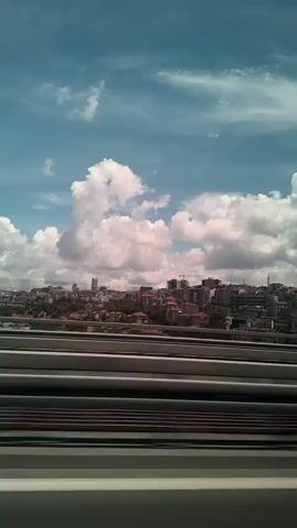 This media file captures a view of Kâğıthane, Turkey, from what appears to be a moving vehicle, possibly a train or bus, looking out over a cityscape. The foreground is dominated by parallel lines of what seem to be train tracks or similar infrastructure, rendered in shades of gray, brown, and white. These lines create a sense of motion and perspective.

Beyond the foreground, the mid-ground reveals a dense urban landscape. Buildings of varying heights and architectural styles, many with reddish-brown tiled roofs, are clustered together on a gently sloping hill. Some taller, more modern structures, including a few skyscrapers and buildings under construction with visible cranes, punctuate the skyline, indicating a mix of old and new development.

The background is a vast expanse of sky filled with dramatic, puffy white clouds against a vibrant blue. The lighting suggests it is daytime, likely midday or early afternoon, with the sun illuminating the clouds and casting shadows on the city. The weather appears to be fair, with a clear sky punctuated by these formations.

There are no people visible in the frame, focusing the attention entirely on the urban environment and the sky. The overall impression is one of a dynamic, developing city seen from a transitional space, perhaps on a journey. The composition, with the repeating lines in the foreground and the sprawling city in the distance, evokes a feeling of travel and observation. There is no visible text in the media file.