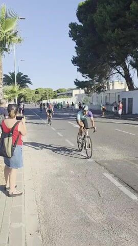 A bicycle race is underway on a sunny day. Cyclists are riding down a paved road, with spectators standing on the sidewalk watching. One spectator in the foreground, a woman wearing a red tank top and a blue denim skirt, is holding up a phone to record the event. The road is marked with white lines, and there are trees and buildings lining the street. Some of the cyclists are wearing helmets and racing attire. The overall atmosphere appears to be energetic and competitive.