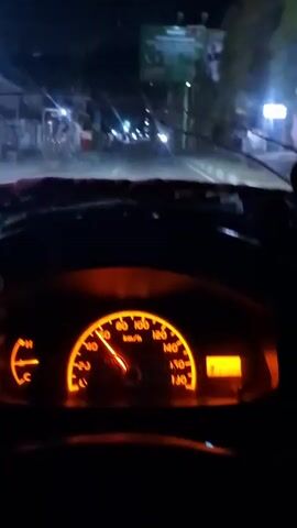 The video is filmed from the driver's perspective inside a vehicle at night. The dashboard, illuminated by an orange glow, displays the speedometer, which indicates the vehicle is traveling at approximately 70 km/h. The fuel gauge is also visible. Through the windshield, the road ahead is lit by the vehicle's headlights, revealing streetlights, buildings, and other vehicles in the distance. The overall scene depicts driving on a road at night.