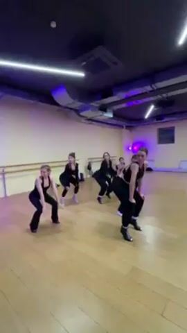 In a wide shot, six women are practicing a dance routine in a dance studio. They are all dressed in black workout attire, including pants, tops, and shoes. The studio has light wooden floors, a mirrored wall, a ballet barre, and yellow walls. The overhead lighting is a mix of fluorescent and purple lights. The women execute dance moves in unison, including bending, reaching, and posing.