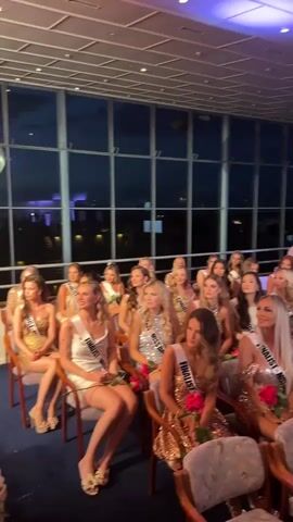 A wide shot shows the finalists of a beauty pageant sitting in chairs and clapping their hands. The women are wearing sparkly dresses and sashes that say “Finalist - Miss…”.  Each woman is also holding a red rose. They are in a large room with large windows in the background that show a twilight scene. There is a white spherical light fixture on the ceiling, along with lighting equipment.  A woman in a pink dress is standing in the back of the room.