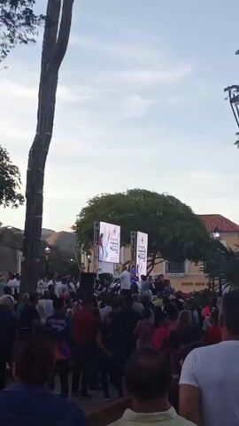 The video shows a large crowd gathered in a public square, looking towards a stage. Two video screens flank the stage, displaying images, likely related to the event. People of various ages and ethnicities are present, dressed in casual clothing. Some individuals appear to be focused on the stage, while others are conversing or looking around. Tall trees and lamp posts are present, adding to the ambiance of the location. A building in the background has "Museo Nueva" written on it. The lighting suggests it might be early evening. The video captures a sense of community and excitement.
