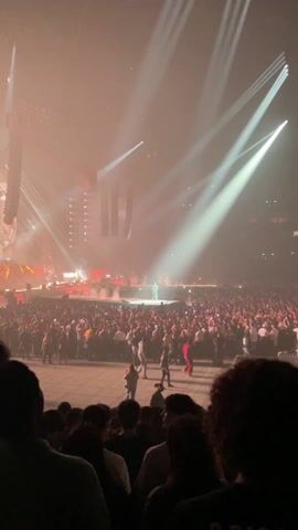 A large crowd of people is gathered in a stadium or large indoor venue for a concert. The stage is visible in the distance, with a performer visible in the center, bathed in bright white spotlights. There are pyrotechnics onstage, with flames shooting up intermittently. Speakers and other equipment are visible overhead. Many people in the audience are holding up their phones, presumably recording the performance. The lighting in the venue shifts colors, including white, green, and pink. The atmosphere appears energetic and lively.
