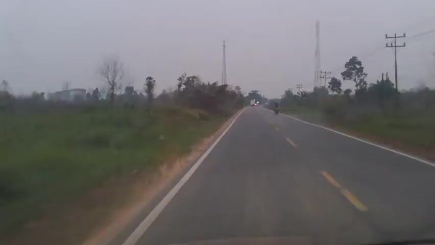 This is a first-person perspective video taken from a moving vehicle on a rural road during daylight hours, likely early morning or late afternoon due to the overcast sky and dim lighting. The road is paved and appears to have a white line marking the shoulder and yellow lines dividing the lanes. To the left of the road, there is a grassy shoulder with some scattered vegetation and trees. In the distance, there are at least two cell phone towers and power lines. There are other vehicles visible on the road, including a motorcycle to the right and what appears to be a car or truck further ahead. The sky is a uniform grey and the overall impression is one of a quiet, somewhat desolate landscape.