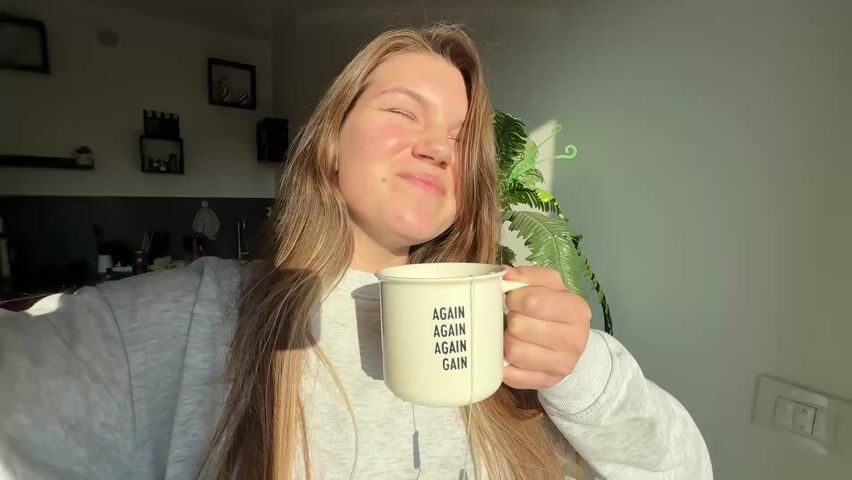 A woman with long brown hair, wearing a gray sweatshirt, is holding a mug with the words "AGAIN AGAIN AGAIN GAIN" printed on it. She is standing indoors, bathed in sunlight. Her head is tilted back and she smiles at the camera. A fern plant is visible behind her, and there are shelves and other items in the background.