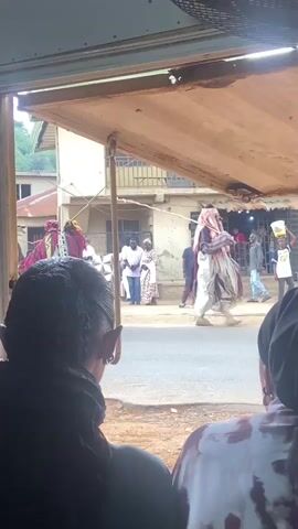 From the interior of a building, a person with braids and another person with a head covering are observing a street scene. The street is filled with people, some in traditional attire and some in contemporary clothing, moving about.  A small dark car is seen driving on the road. Buildings are visible in the background, along with a hillside covered with trees.  The perspective is through a window with a partially open awning. The sound of drumming can be heard.