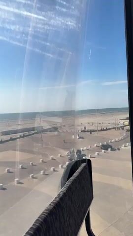 The video shows a view from inside a Ferris wheel cabin as it ascends. Outside the cabin's windows, a sunny blue sky is visible. In the distance, a body of water, likely the ocean, meets the horizon. The foreground features the interior of the cabin, including a seat with a patterned fabric, part of the cabin's structure, and a window frame. As the cabin moves, the view changes, revealing the intricate white steel structure of the Ferris wheel itself, with a ladder system visible. Below, a paved area with parked cars and white bollards can be seen. The overall impression is that of an amusement park or a similar recreational area.