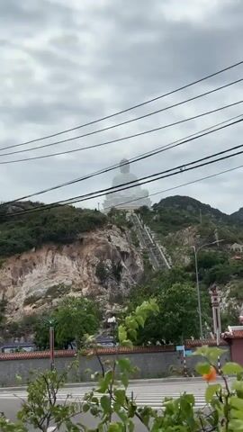 This is a vertical video of a large white Buddha statue seated on a hilltop. The statue is visible in the background, elevated above a lush green hillside. In the foreground, there is a paved road with a pedestrian crossing. To the right, a traditional Vietnamese temple gate with ornate red and gold detailing is partially visible. A steep set of stairs leads up the hillside towards the Buddha statue, and several power lines are strung across the top of the frame. The sky is overcast and cloudy.