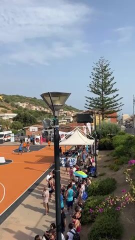 The video shows a basketball court with an orange surface, set in a hilly, urban environment. Several people are playing basketball on the court. There is a crowd of spectators watching the game, seated and standing along the edge of the court. Tents are set up near the court, and there are buildings and trees in the background. The sky is blue with some clouds. The lighting suggests it is daytime, likely outdoors.