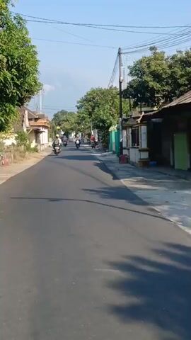 The video is a first-person perspective recording of riding a motorcycle down a paved street in a village. The street is lined with houses and trees. Several other motorcycles are visible on the road, some passing by, others in the distance. The weather appears to be clear and sunny. The recording starts with a view of the street ahead, then shows other motorcyclists, and continues down the road with buildings and foliage on either side.