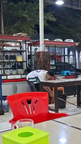 The video shows a young man wearing a white t-shirt with green and black stripes, and dark pants, sweeping the floor with a broom. He is in an outdoor setting, likely a food stall or restaurant, as indicated by the tables, chairs, and display cases with food items visible in the background. Another person, a young man in a blue shirt, is visible in the background on the right side, holding a bottle of water and a blue object. The scene appears to be at dusk or nighttime, with artificial lighting illuminating the area. There are red chairs and tables in the foreground, and a green tissue box is on a table in the immediate foreground. The overall impression is one of a casual eatery being cleaned up at the end of the day.