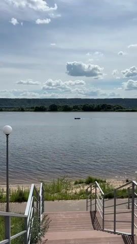 This is a vertical video clip of a river landscape on a cloudy day. The foreground features a wooden staircase with metal railings leading down towards a sandy shore. To the left of the stairs, a modern lamp post with a white spherical light is visible. The middle ground is dominated by a wide, calm river, its surface rippling gently. In the distance, a small dark boat with two people on board is moving across the water, leaving a visible wake. The background showcases a lush, tree-covered hillside stretching across the horizon. The sky is partly cloudy with large, white cumulus clouds interspersed with patches of blue sky. The overall mood is peaceful and serene, suggesting a pleasant, perhaps warm, day. The time of day appears to be daytime, likely mid-afternoon given the lighting. The location is identified as Zvenigovo, Russia. There is no visible text in the media file.