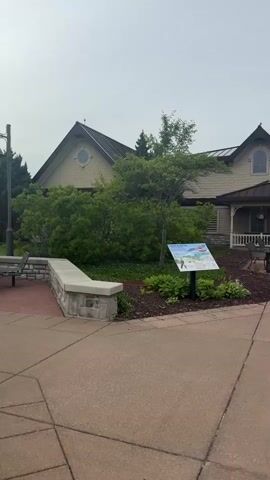 The video captures an exterior view of a building with a light-colored facade and a dark, multi-gabled roof. A covered porch with white railings runs along the front of the building, and there are red double doors at the entrance. In front of the building, there is a paved walkway and a landscaped area with a sign, mulch, plants, and a trash receptacle. The sky is overcast.