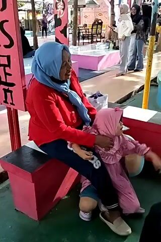 An adult wearing a blue hijab and a red long-sleeved shirt is sitting on a pink bench. They are holding a child dressed in a pink hijab and a pink dress. The child is sitting on the adult's lap, facing to the right. The adult's left arm is around the child. The child's right arm is extended outwards. The adult's right hand is resting on the child's leg. The adult is looking towards the left. In the background, several other people are visible, some walking and some standing. The environment appears to be outdoors, possibly a market or a public space with colorful structures and a green ground surface. There is a pink sign with black writing to the left of the adult and child.