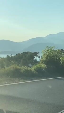 The video shows a view from a car traveling on a road. The road has a white line on the right side and a reflective barrier running along the left side of the road. In the background, there are lush green trees and hills, with a modern white building visible among the trees on the right side of the frame. The sky is clear and blue, suggesting a sunny day. The car appears to be moving at a moderate speed, as indicated by the slight blur of the scenery. The overall impression is that of a scenic drive through a coastal or mountainous region.
