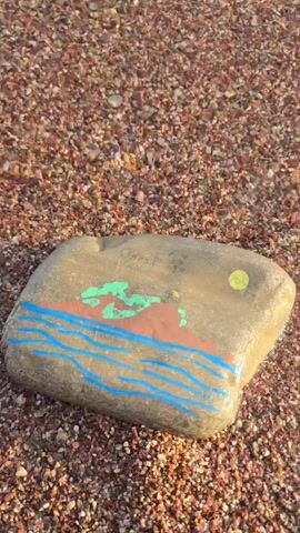 The video begins with a close-up of a painted rock on a pebble beach. The rock has a simple drawing of a seascape, with blue waves, a red-orange landmass, and green foliage under a yellow sun. The scene then pans out to show the shallow, clear water gently lapping at the shore, revealing the colorful pebbles beneath the surface. The camera continues to move towards the sea, showing a wider view of the bay. In the distance, there are several boats, including a white sailboat and a larger white vessel. A small island with some rocky outcrops is visible in the center of the bay. The sky is clear with a few wispy clouds, suggesting it is a pleasant day. The light indicates it is likely late afternoon or early morning. The overall mood is peaceful and serene, typical of a beach setting. There is some faint, illegible writing on the painted rock.