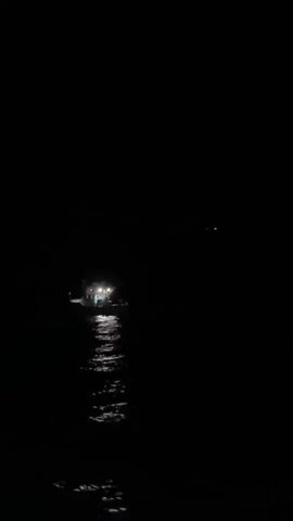 The video begins with a shot of a dark body of water at night. A distant boat with lights on is visible in the center of the frame. The lights reflect on the water, creating a shimmering effect. The boat appears to be stationary. The surrounding environment is completely dark, with no other discernible features. The camera remains still throughout the short clip.