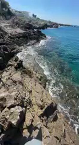 The video captures a first-person perspective of a scenic rocky coastline on a bright, sunny day. The initial shot shows the videographer's legs, clad in light-colored pants and shoes, resting on dark, jagged rocks that descend into crystal-clear, turquoise water. Small waves gently break against the shore, creating white foam.

The camera then pans up to reveal a stunning expanse of the sea, its vibrant blue-green hues contrasting with the rugged, dark grey rocks. The horizon is visible under a brilliant, cloudless blue sky, with the sun shining intensely, causing a lens flare in some frames. The coastline stretches into the distance, marked by more rocky formations and sparse vegetation. Another pan shows a section of rocks covered in green algae or moss, indicating the water line. The video concludes with a wide view of the serene seascape, emphasizing the clear water and natural beauty of the rocky shore. The setting appears tranquil and idyllic. No visible text is present. The location is Nardò, Italy.