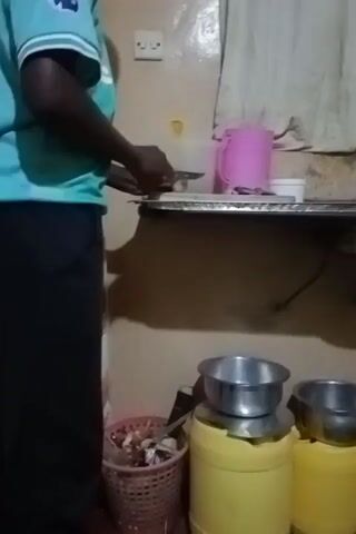 A person wearing a teal shirt and black pants is standing in a kitchen area. They are holding a utensil and appearing to be preparing food on a metallic counter. On the counter, there are several items including a pink container, a white container, and what appear to be onions. Below the counter, a wicker trash bin is visible, filled with miscellaneous items. To the right, two yellow containers are stacked, with metal bowls placed on top of them. The walls are a light brown color, and a white electrical outlet is mounted on the wall. The lighting is dim.