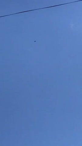 The video shows a blue sky with a single, distant, dark triangular object in the center of the frame. Later, another dark shape appears above it, and a bird flies below the triangular object. A power line is visible at the top left of the frame.