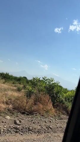 The video begins with a wide shot of a bright, clear blue sky with a few wispy clouds. In the foreground, lush green foliage with dry, brown grasses dominates the lower portion of the frame. In the middle ground, more trees and bushes are visible, and in the far distance, two majestic, snow-capped mountains rise above the horizon. The scene then transitions to a view from inside a vehicle, looking out the side window. The view shows a dirt road with rough terrain and more greenery. The video then shifts to a view from inside the vehicle, looking through the windshield. The camera captures a long, dusty dirt road stretching out ahead, flanked by sparse vegetation and trees. The sun is bright, casting long shadows and creating a hazy effect on the distant landscape. The dashboard of the vehicle is visible in the lower part of the frame, including a steering wheel and part of the radio.
