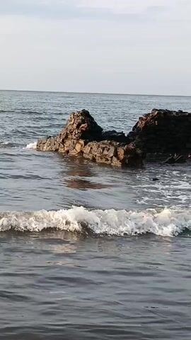 Here's a concise description of the video:

The video shows a view of the ocean, with small waves gently rolling towards a rocky outcropping. The rocks are brownish-black in color and appear to be partially submerged in the water. The ocean extends to the horizon under a mostly clear sky.