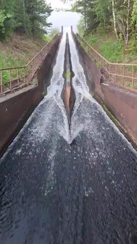 The video shows a perspective from above looking down a concrete spillway with water flowing down its center. The spillway is flanked by metal railings and earthen banks covered in grass and trees. The water is turbulent, creating white foam as it cascades down the incline. In the distance, a body of water, likely a lake or reservoir, is visible, surrounded by a tree line under an overcast sky.