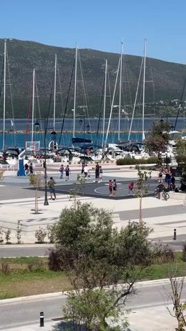The video shows a basketball court in a marina with several sailboats docked in the background. People are playing basketball on the court. Some are actively playing, while others are watching. There are also people walking and riding bikes in the surrounding area. The weather appears to be sunny and warm.
