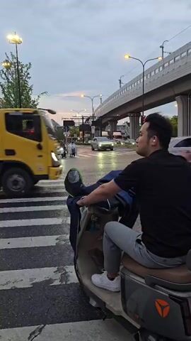 A person sits on a scooter at a crosswalk. The person is wearing a black shirt and grey pants. They are looking to the right. A yellow truck is driving across the crosswalk in the background. A white car is also driving across the crosswalk. The sky is overcast.