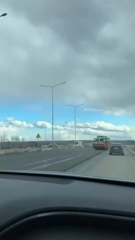 The video captures a forward-facing view from inside a moving car on a multi-lane road during daytime. The sky is partly cloudy with visible patches of blue. In the rightmost lane, road construction is underway. An orange road roller, with the brand "HAMM" visible on its side, is prominent in the foreground, slowly moving along the road surface. Behind it, a yellow road roller and an asphalt paver are visible, indicating road paving activities. Traffic cones mark the construction zone. Streetlights line the road, and other vehicles, including cars and a white bus, are seen in adjacent lanes and in the distance. The dashboard of the recording vehicle is visible at the bottom of the frame. The scene depicts an active road construction or maintenance site.