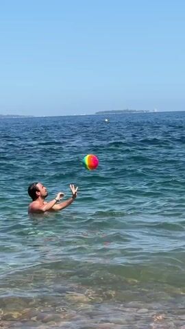 A man in the water is throwing a rainbow colored ball to another man. The sun is shining and the sky is clear. The water is blue and clear. The men are playing catch in the water.