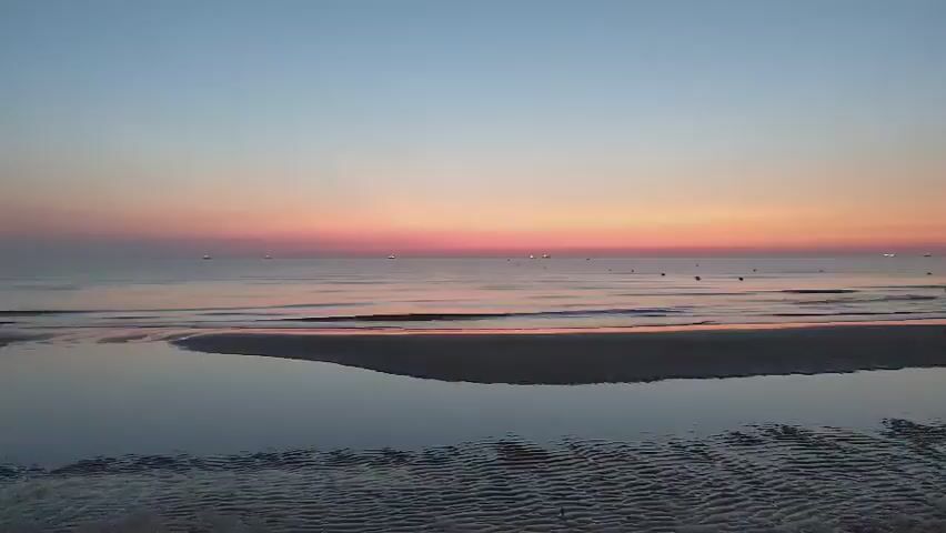 A serene beach scene unfolds at twilight. The sky transitions through shades of pale blue, soft pink, and orange, creating a beautiful gradient above a calm sea. Gentle waves lap at the shore, reflecting the colors of the sky. A small inflatable boat with an outboard motor rests partially on the wet sand, its reflection visible in the shallow pools of water left by the receding tide. In the distance, small lights dot the horizon, possibly indicating other boats or buoys. The overall atmosphere is peaceful and tranquil.