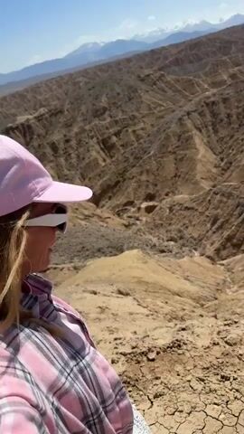 A person, seen from the side and slightly behind, stands on a dry, earthy ridge, looking out over a vast, arid landscape. The individual wears a pink baseball cap, white sunglasses, and a pink plaid long-sleeved shirt, with a white woven bag visible on their right side.

The scene reveals a deeply eroded canyon or valley, characterized by numerous layered, reddish-brown hills and slopes. The ground beneath the person's feet is dry, cracked earth and loose soil, with sparse, low-lying vegetation. In the far distance, majestic snow-capped mountains stretch across the horizon under a clear, bright blue sky. The lighting suggests it is daytime, likely a sunny morning or afternoon. The camera slowly pans, showcasing the immense scale of the mountainous terrain. The location is Bolshevik, Kyrgyzstan, which aligns with the Central Asian mountainous, desert-like features of the landscape. There is no visible text.