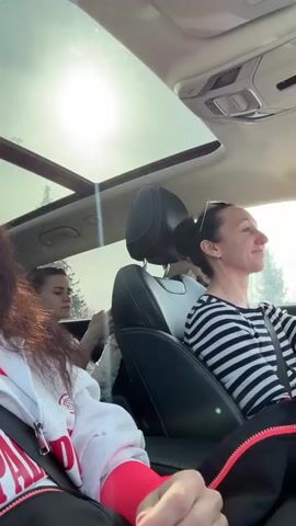 A bright, sunny video captures three women inside a car, seemingly on a road trip in the Gornyak area of Russia. The car features a panoramic sunroof, through which intense sunlight streams into the cabin.

In the foreground, a woman with long, dark, wavy hair is taking a selfie or recording a video. She wears a light grey zip-up hoodie over a bright red long-sleeved top, with a black bag strap across her chest and a seatbelt fastened. She looks directly at the camera with a relaxed, pleasant smile. Partially visible red lettering on her hoodie reads "PA..."

In the midground, in the backseat on the left, another woman with dark hair tied back is partially visible. She appears to be focused on something in her hands or adjusting her hair.

The driver, positioned on the right side of the vehicle (indicating a left-hand drive car), has dark hair pulled back and sunglasses resting on her head. She wears a black and white striped long-sleeved shirt. She looks out the driver's side window with a calm, pleasant smile, enjoying the drive.

All three women are wearing seatbelts. The background visible through the car windows and sunroof shows clear skies and some trees. The overall mood is cheerful and relaxed, with music playing in the background.