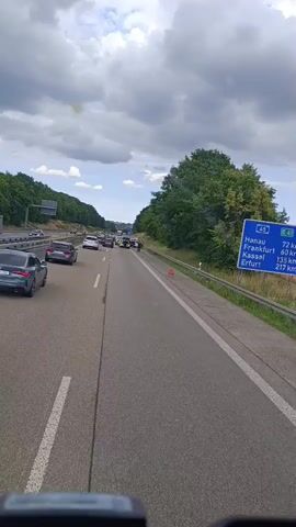 A traffic jam is seen on a highway under a cloudy sky. Cars are stopped in multiple lanes. A hazard triangle is visible on the side of the road, indicating a potential accident or breakdown. A signpost displays destinations such as Hanau, Frankfurt, Kassel, and Erfurt, along with distances. The perspective is from inside a vehicle, looking forward.