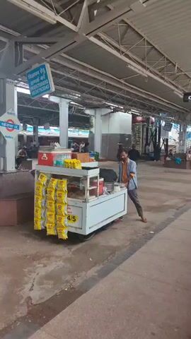 The video shows a train station platform. A food cart is visible on the left side of the frame, stocked with snacks. A man stands near the cart. Several people are sitting on benches further down the platform. A train is partially visible on the right side of the frame. The platform is covered by a roof supported by metal beams. The ground appears to be concrete.