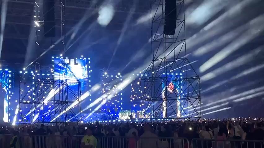 This is a wide shot of a large outdoor concert or festival with a massive crowd gathered. The stage is elaborately set up with numerous lights and large screens displaying performers. Smoke machines are creating a hazy atmosphere, and beams of light crisscross the sky. The audience is visible in the foreground, standing behind a barricade. The overall impression is one of a vibrant and energetic event.