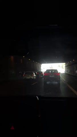 The video shows a first-person perspective from inside a car as it exits a dark tunnel and enters a brightly lit highway. Multiple vehicles are visible on the highway, including a black SUV in front of the car and several other cars in the distance. Trees and greenery line the sides of the highway. The camera angle is looking forward through the windshield and shows the car's dashboard and rearview mirror. There is a brief audio clip of a person speaking in Spanish, which translates to "that it leaned on that I started to feel how the seatbelt pulled me back."