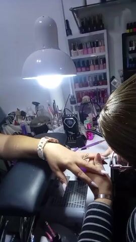 Here's a description of the video:

The video shows a nail technician working on a client's nails. The client is wearing a white watch on their left wrist and has long, white stiletto nails. The technician is wearing glasses and a striped top. They are using a tool to work on the client's cuticle area, possibly pushing back or cleaning the area.

The setting appears to be a nail salon or a nail technician's workspace. There's a bright white lamp shining down on the workspace. A shelf behind the technician is filled with various bottles of nail polish. There are also other tools and equipment on the table, including brushes, containers, and a nail lamp.