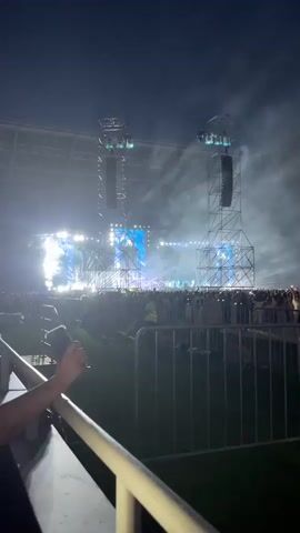 The video shows a nighttime outdoor concert at a stadium. The stage is set up with large speaker towers and a big screen. There is a lot of smoke and bright lights illuminating the stage and the audience. The audience is visible as a large crowd of people gathered in front of the stage, with some people holding up their phones. The foreground shows a metal barrier with a green grassy area behind it. A person's arm and hand holding a phone are visible in the lower left corner of the frame. There are also a few people in yellow safety vests visible within the crowd behind the barrier. The sky is dark, indicating it is nighttime.