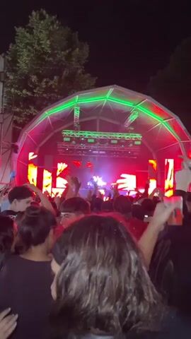 A nighttime music event is depicted from within a large crowd facing a brightly lit stage. The stage is covered by a tent-like structure illuminated with green lights along its framework. A DJ is visible at the center of the stage, operating equipment. Behind the DJ, a large LED screen displays dynamic abstract graphics of glowing red and orange hands. Red and white stage lights are active on the stage itself. Numerous individuals in the foreground and mid-ground of the crowd have their hands raised in the air, engaging with the performance.
